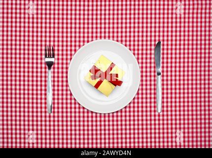 Surprise meal, a gift wrapped present box on a round white plate with knife and fork from above on a red gingham tablecloth Stock Photo