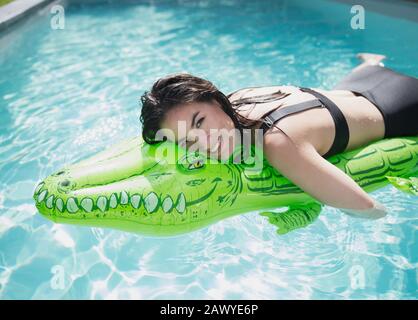 Portrait happy, carefree woman floating on inflatable alligator raft in sunny summer swimming pool Stock Photo