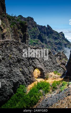 Small pedestrian path at the top of Madeira mountain range above clouds. The path is surrounded by green grass and goes through round hole in volcanic Stock Photo
