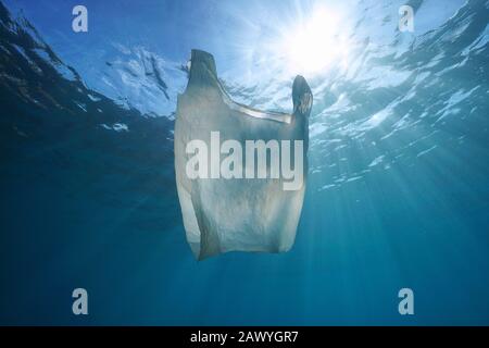 Plastic pollution in ocean, a white plastic bag drifts underwater with sunlight through water surface Stock Photo