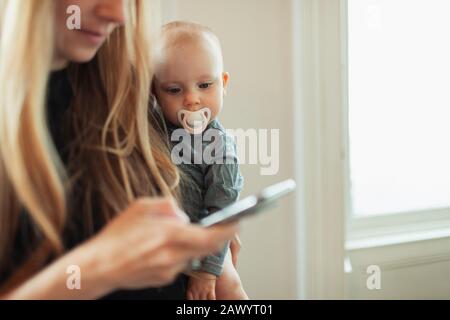 Curious baby girl with pacifier watching mother using smart phone Stock Photo