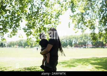 Portrait happy father with long braids carrying son in sunny park Stock Photo