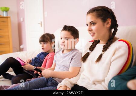 Boy with Down Syndrome watching TV on sofa with sisters Stock Photo