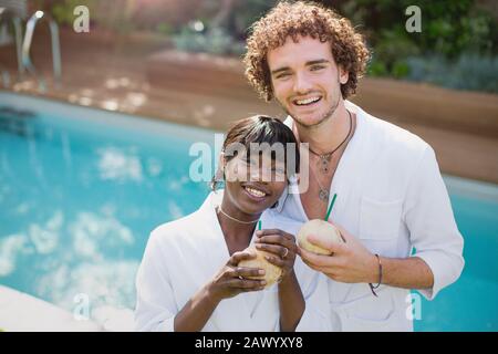 Portrait happy young couple in bathrobes drinking from coconuts at poolside Stock Photo