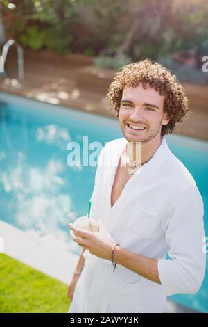 Portrait happy young man in bathrobe drinking from coconut at poolside Stock Photo