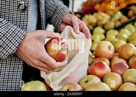 closeup of a man shopping at a greengrocer putting some red apples in a textile reusable mesh bag, as a measure to reduce plastic pollution Stock Photo
