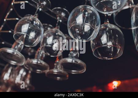 Closeup many upside down empty clear transparent crystal upturned wine glasses hanging in straight rows on brown wooden shelf, rack, showcase, stainle Stock Photo