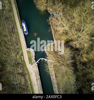 English canal scene with a  narrowboat barge, lock and towpath surrounded by winter trees on a bright February day. Vertical drone view. Stock Photo