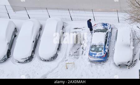 Bucharest, Romania - February 6, 2020: Man clears snow from his car using a brush, on a row with parked car covered by a fresh layer of snow. High ang Stock Photo