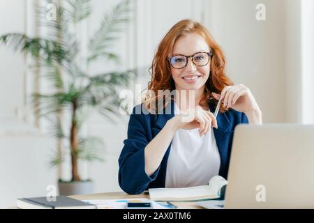 Portrait of happy redhaired woman employee in optical glasses, has satisfied expression, works with modern gadgets, waits for meeting with colleague, Stock Photo