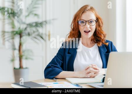 Surprised ginger female employee has job, develops new business srategy, poses in front of opened laptop computer, poses in workspace against office i Stock Photo