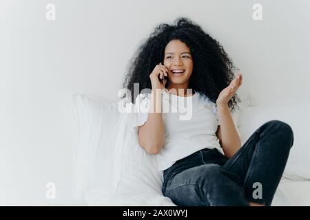 People, technology, communication and rest concept. Glad curly haired ethnic lady has telephone talk, gestures and laughs with joy, looks somewhere as Stock Photo