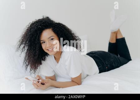 Indoor shot of cheerful millennial woman with Afro hairstyle, lies on stomach in comfortable bed, chats online, checks email box, wears white t shirt, Stock Photo