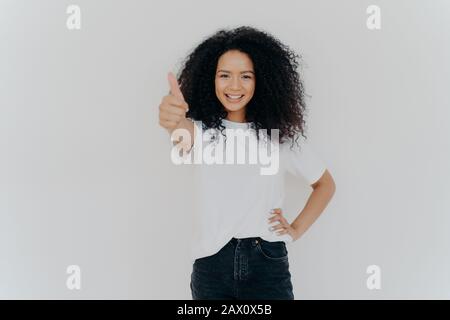 Photo of happy curly woman with crisp hair raises thumb up, gives approval, says sounds good, makes supportive gesture, keeps other hand on waist, dre Stock Photo