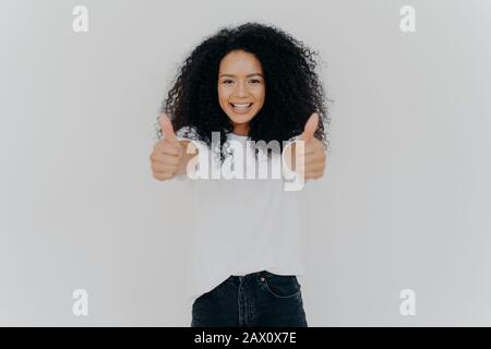 Happy young woman with Afro hairstyle, shows thumbs up, accepts or agrees awesome plan, gives her approval, smiles broadly, stands indoor, says excell Stock Photo