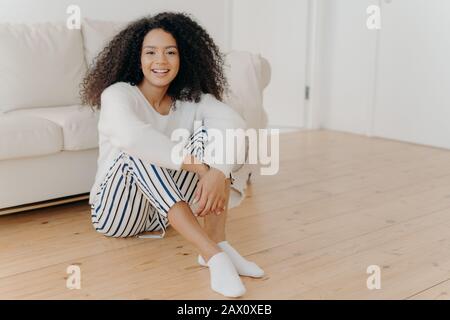 Indoor shot of restful happy African American woman sits on floor in living room, white sofa in background, wears fashionable clothes and socks, expre Stock Photo