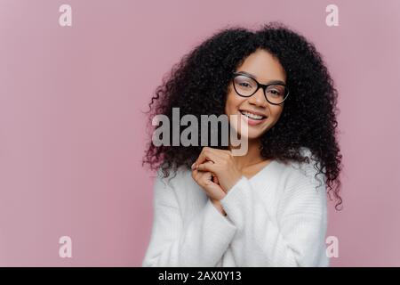 Portrait of happy positive African American woman with curly bushy hair, keeps hands together, has pleased facial expression, wears spectacles and whi Stock Photo