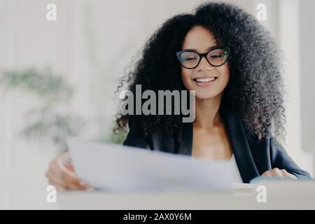 Happy Afro American woman with curly hairstyle, looks through documents, prepares financial report, wears transparent glasses and formal suit, poses i Stock Photo