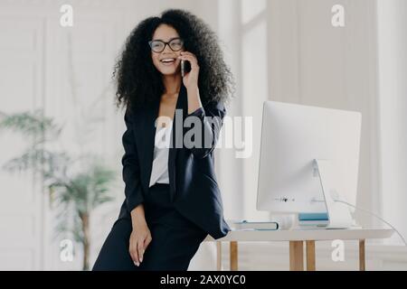 Image of satisfied dark skinned businesswoman leans at white table with computer, has break after work, poses in her cabinet, speaks via cellular, wea Stock Photo