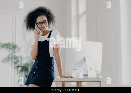 Pleasant looking African American female has telephone conversation, asks for advice before job interview, dressed in stylish clothes, poses near work Stock Photo