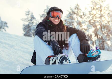 Stock photo of a young girl snowboarder is smiling and sitting on the snow of the mountain Stock Photo