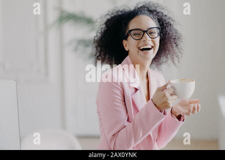Overjoyed curly haired woman laughs happily while drinks hot coffee or latte, wears transparent spectacles, has fun during break in office, shows whit Stock Photo