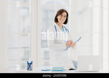 Skilled female therapist or cardiologist writes information at clipboard, ready to consult and cure patients, wears white coat, poses at hospital offi Stock Photo