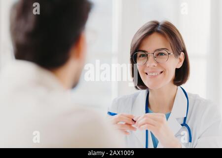 Smiling brunette woman doctor in round glasses, wears white coat, has conversation with patient, happy to help other people, wears white coat uniform, Stock Photo