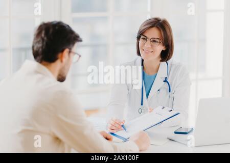 Young female doctor consults patient, shows information and prescription in clipboard, talk about medical checkup, pose at clinic, explains health ins Stock Photo