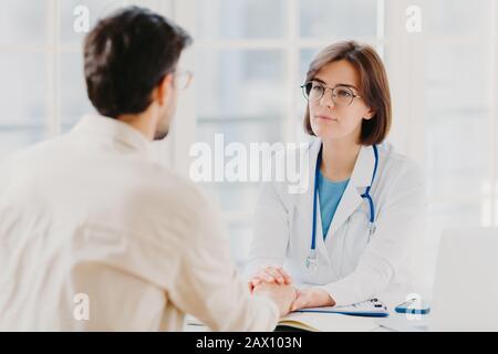 Friendly female doctor tries to support patient, holds his hands, gives useful consultation and explains medical information, makes diagnostic examini Stock Photo