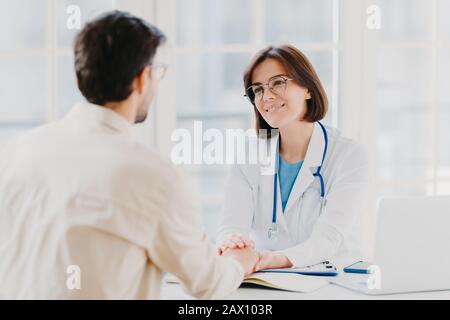 Confident female doctor holds hands of ill patient, persuades everything will be alright, dressed in white medical gown, gives advice, pose in hospial Stock Photo