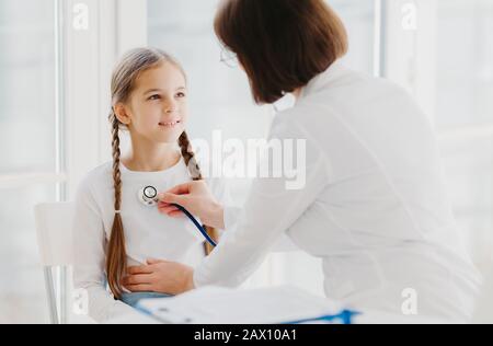 Caring female pediatrician listens childs lungs or heart with phonendoscope. Little girl comes on examination in private clinic, has every year doctor Stock Photo