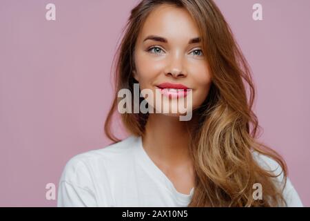 Horizontal cropped image of pleasant looking female model has tender smile, wears minimal makeup, has long wavy hair, looks at camera with satisfactio Stock Photo