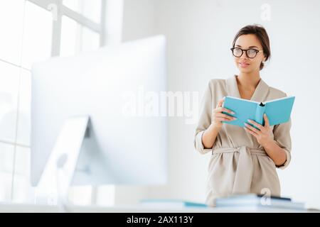 Young dark haired woman in elegant wear holds blue notepad focused into monitor, ponders about business strategy for eneterprise, wears transparent ey Stock Photo