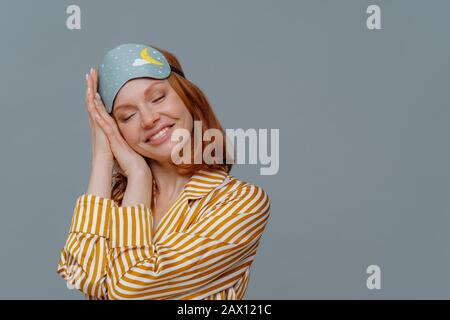 Satisfied ginger woman has nap, sees pleasant dreams, tilts head on palms, smiles gently with closed eyes, wears sleep mask, dressed in striped pajama Stock Photo
