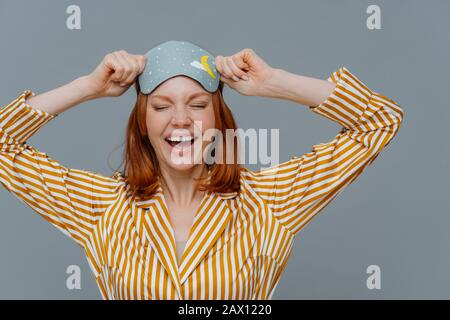 Overjoyed red haired Caucasian woman laughs positively, wears blindfold and yellow striped pajama, expresses good emotions, isolated on grey backgroun Stock Photo