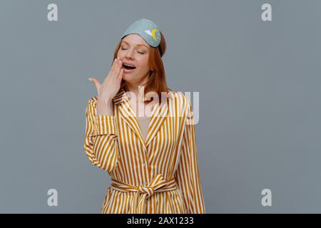 Fatigue sleepy red head woman yawns with opened mouth, wears domestic striped robe and blindfold on head, wants to sleep, stands against grey backgrou Stock Photo