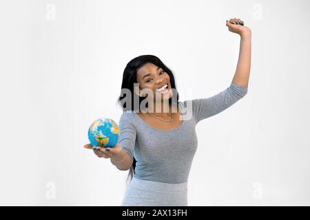 Young african woman standing isolated on white wall holding litlle earth globe, looking camera, smiling with one hand up. Travel, geography concept. Stock Photo