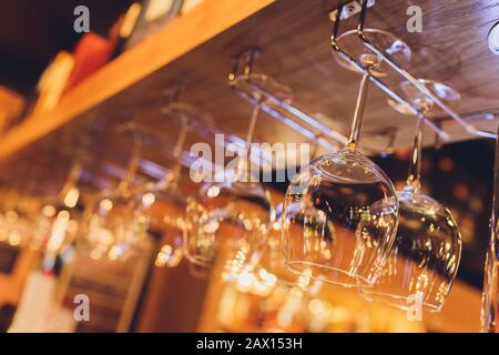 Closeup many upside down empty clear transparent crystal upturned wine glasses hanging in straight rows on brown wooden shelf, rack, showcase, stainle Stock Photo