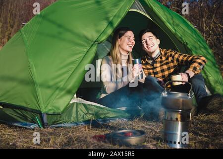 happy young couple in love with nature smiling, drinking tea and coffee in tent near a fire. outdoors activity for young families Stock Photo