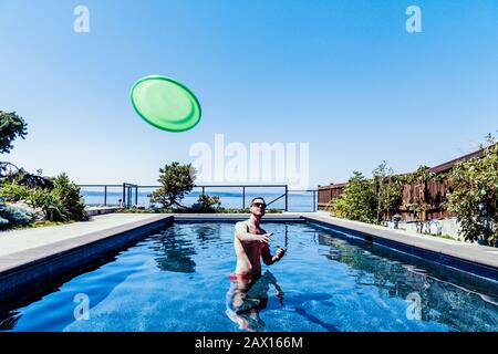 Man in standing in backyard swimming pool on a hot summer day tossing a green frisbee flying disc toward the viewer. Stock Photo