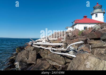 Sun bleached driftwood resting on reinforced rock sea wall under the Alki Point Lighthouse in West Seattle, Washington, mid day blue sky. Stock Photo