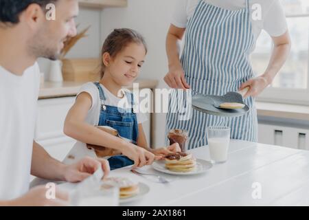 Unrecognizable mommy brings delicious pancakes to table, prepares breakfast for family. Cheerful girl adds melted chocolate to dessert, enjoys time wi Stock Photo
