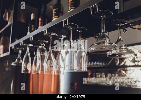 Closeup many upside down empty clear transparent crystal upturned wine glasses hanging in straight rows on brown wooden shelf, rack, showcase, stainle Stock Photo
