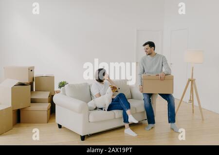 Positive woman and man pose in empty spacious room during relocation day, husband carries cardboard boxes with belongings, wife has telephone conversa Stock Photo