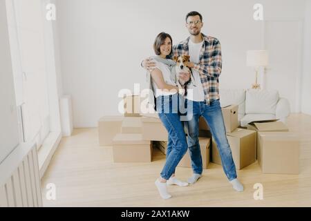 Young couple in love stand closely to each other, embrace and holds favourite dog, pose in empty room, surrounded with cardboard boxes, make family po Stock Photo