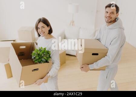 Positive female and male property owners pose with personal belongings in carton boxes, relocate to own apartment, smile pleasantly, pose in white liv Stock Photo