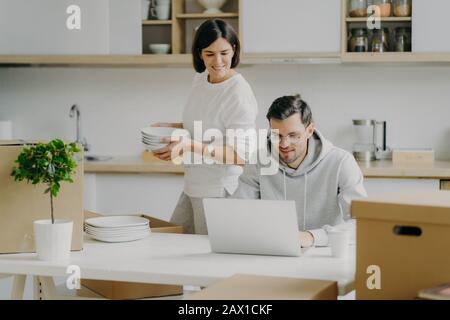 Young brunette woman unpacks boxes with personal belongings, places plates in cupboard, looks attentively at laptop computer where husband works. Fami Stock Photo