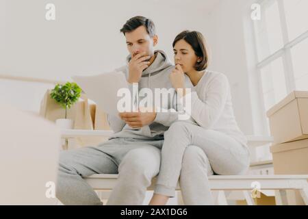 Photo of upset woman and man have unpaid domestic bills, hold papers, sit in spacious room, manage finances, review bank account, have to pay taxes, r Stock Photo
