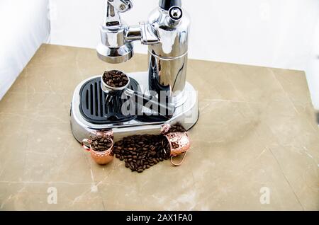 Spilled coffee beans in front of an espresso machine, placed on it with a lever of beans. Metal cups in copper color. Stock Photo
